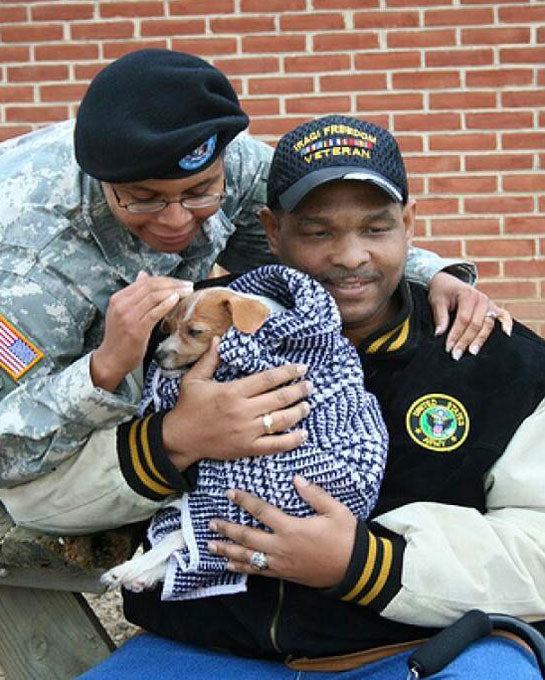 U.S. Veteran man sitting holding small puppy in blue and white blanket with woman in military fatigues putting arm around man's shoulder looking at puppy