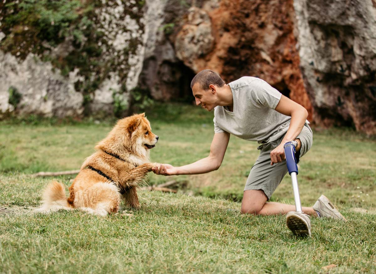 Man with prosthetic leg kneeling in field of grass, shaking hands with an orange dog